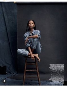 a woman sitting on top of a wooden stool in front of a black backdrop with her legs crossed