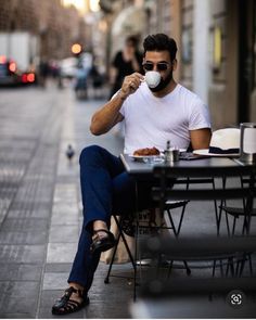 a man sitting at an outdoor table drinking from a cup while holding a coffee mug