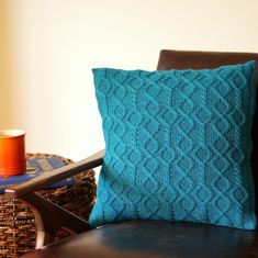 a blue knitted pillow sitting on top of a brown chair next to a cup