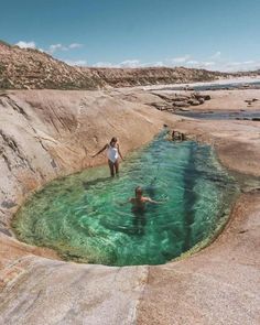 two people are swimming in the water near some rocks and sand, while another person is standing on one side of the pool