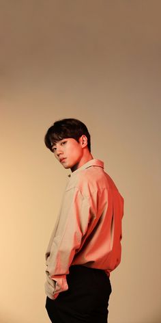 a young man standing in front of a white wall wearing an orange shirt and black pants