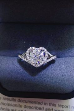 an engagement ring with a heart shaped center surrounded by round brilliant cut diamonds in a blue velvet box