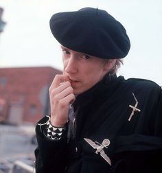 a young man wearing a black hat and holding his finger to his mouth while standing in front of a building