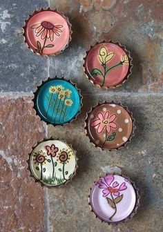 four bottle caps with flowers painted on them sitting on a stone floor next to a brick wall