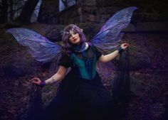 a woman dressed as a fairy with purple hair and blue wings sitting in the woods