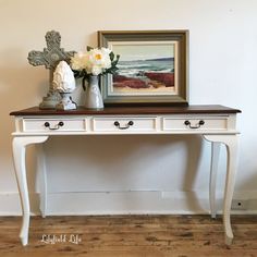 a white table with two drawers and flowers on it next to a framed painting in a room