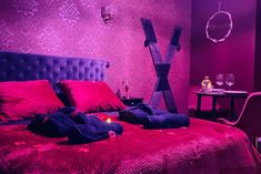 a bed with red sheets and pillows in a purple room next to a table with two wine glasses on it