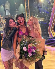 three women standing next to each other in front of a stage with flowers on it