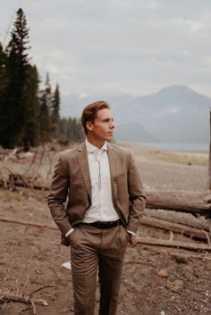 a man wearing a suit and tie standing in front of a tree trunk with mountains in the background