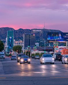 cars are driving down the street in front of tall buildings and mountains at dusk with lights on