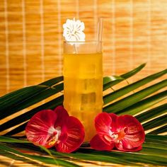 two red flowers sitting next to a drink on top of a bamboo table with green leaves