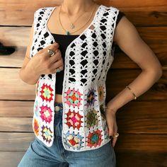 a woman standing in front of a wooden wall wearing a crocheted vest and jeans