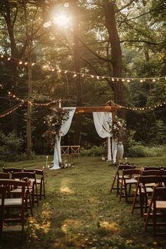 A beautiful garden wedding setup with a wooden arch draped in white fabric and adorned with floral arrangements, surrounded by string lights and wooden chairs under tall trees. Hers And Hers Wedding Ideas, Forest Backyard Wedding, Wedding In The Garden Decoration, Outdoor Wedding Lighting Ideas Backyards, Wedding By Pond Ideas, Backyard Ceremony Decor, Backyard Budget Wedding, Diy Outdoor Wedding Decor, November Wedding Outdoor