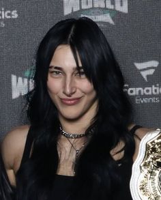 a woman with long black hair holding a gold and white wrestling belt in front of a wall