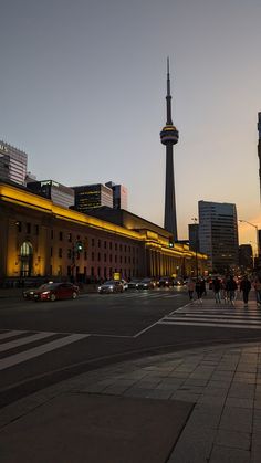 people are walking on the street in front of some tall buildings at dusk with traffic passing by