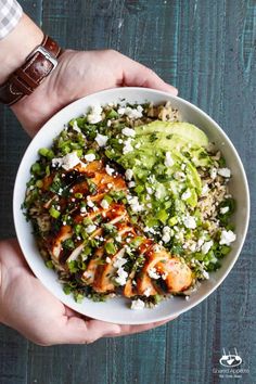 two hands holding a white bowl filled with chicken and rice, guacamole on the side