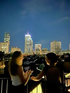 two people are standing on a balcony looking at the city lights and skyscrapers in the distance