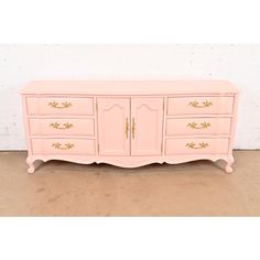 an antique pink dresser with gold trimmings on the doors and drawers, against a white brick wall