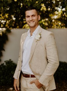 a man in a tan suit and white shirt smiles at the camera with his hands in his pockets