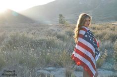 a woman standing in a field with an american flag draped over her shoulders and looking at the camera