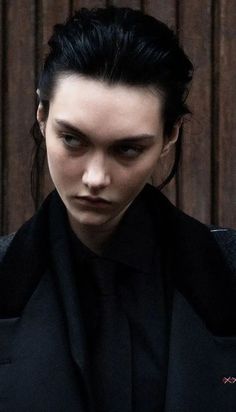 a woman with black hair wearing a dark suit and looking off to the side while standing in front of a wooden wall