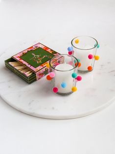 two candles on a marble plate next to a book and some colored pom - poms