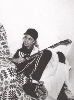 a man sitting on top of a bed with a guitar in his hand and wearing a beanie