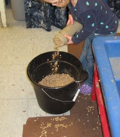 a young child pouring sand into a black bucket on top of a floor next to a blue bin