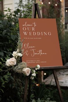 a wedding sign with flowers on it in front of some greenery and string lights