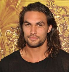 a man with long hair and beard standing in front of a golden wall wearing a black shirt