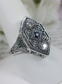 Blue Sapphire & White Cubic Zirconia (CZ) Sterling Silver RingCharlotte Design#D231 Resurrected from the Art Deco movement, this gorgeous Art Deco reproduction filigree ring is crafted in sterling silver. This lovely filigree ring is set with a stunning quality 2mm center round-cut simulated blue sapphire gemstone. Two small white cubic zirconia gemstones also grace the face of the ring, one near each point, each set in the center of a tiny heart. There are 2 filigree accent flowers that grace t Silver Rings With Intricate Cubic Zirconia Design, Antique Silver Sapphire Ring With Diamond Accents, Silver Sapphire Ring With Intricate Design For Formal Occasions, Silver Filigree Ring With Intricate Design For Formal Occasions, Formal Silver Filigree Ring With Intricate Design, Formal Silver Sapphire Ring With Intricate Design, Art Deco Sterling Silver Filigree Ring With Diamond Cut, Silver Diamond Cut Filigree Ring In Fine Jewelry, Silver Sterling Filigree Ring With Intricate Design