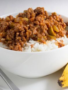 a bowl filled with rice and meat next to a banana on a white tablecloth