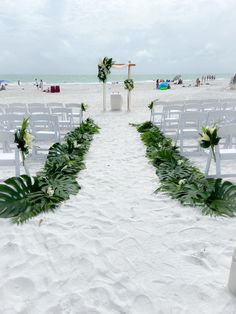 an outdoor ceremony setup on the beach with white chairs and greenery lined up along the aisle