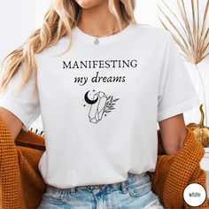 Embrace the power of manifestation with our 'Manifesting My Dreams' Crystal Shirt. Designed with witchy, mystical vibes, this tee blends the energy of crystals with the Law of Attraction. Whether you're manifesting your desires or drawn to spiritual practices, this shirt offers both style and inspiration. Perfect for dreamers, spiritual seekers, or crystal lovers, it also makes a thoughtful gift. Would you prefer this design on a Sweatshirt? Have a look at our other listings here:  https://raidh Spiritual Tee Shirts, Cheap Spiritual Graphic Print T-shirt, Crystal Shirt, Power Of Manifestation, Boho Frame, Spiritual Manifestation, The Law Of Attraction, Spiritual Practices, Law Of Attraction