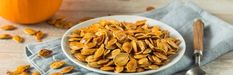 a white bowl filled with pumpkin seeds on top of a blue napkin next to a silver spoon