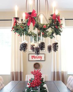 a dining room table decorated for christmas with pine cones and holly wreaths hanging from the chandelier