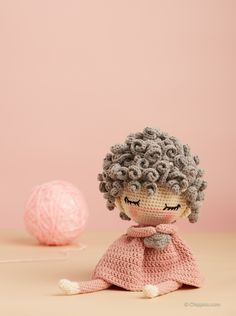 a crocheted doll sitting next to a ball of yarn on a table with a pink background