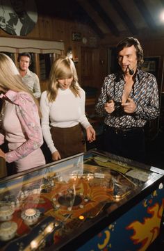 three people standing around a pinball machine in a room with other people looking at it