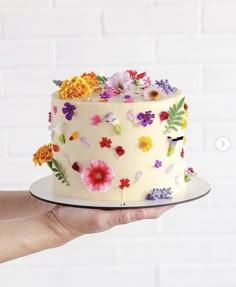 a hand holding a white cake with flowers on the frosting and icing, in front of a brick wall