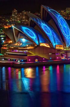 the sydney opera is lit up in blue and purple lights at night, with city lights reflected on the water