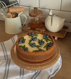 a blueberry cheesecake on a wooden platter next to a tea pot and other kitchen accessories