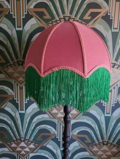 a pink and green table lamp with fringes on the base against a wallpapered background