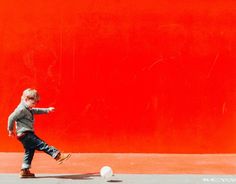 a young boy kicking a soccer ball in front of a red wall