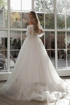 a woman in a white wedding dress standing by a large window looking at the outside