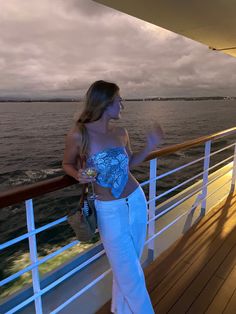 a beautiful young woman standing on top of a wooden deck next to the ocean wearing white pants