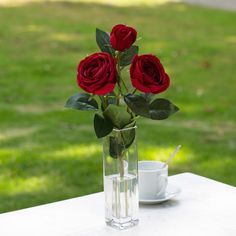 three red roses in a vase on a table with a coffee cup and saucer