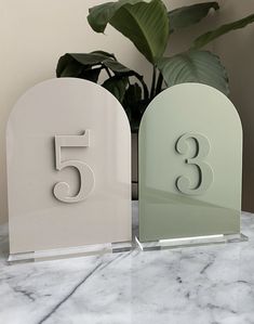 two white and one green glass table numbers sitting on a marble surface next to a potted plant