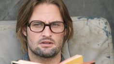 a man with glasses reading a book while sitting in a chair and looking at the camera