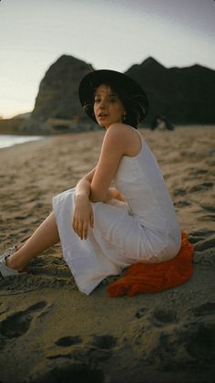 a woman sitting on top of a sandy beach next to the ocean wearing a hat