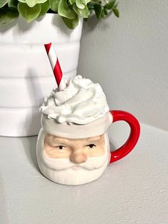 a ceramic santa mug with a candy cane sticking out of it's top, sitting on a table next to a potted plant